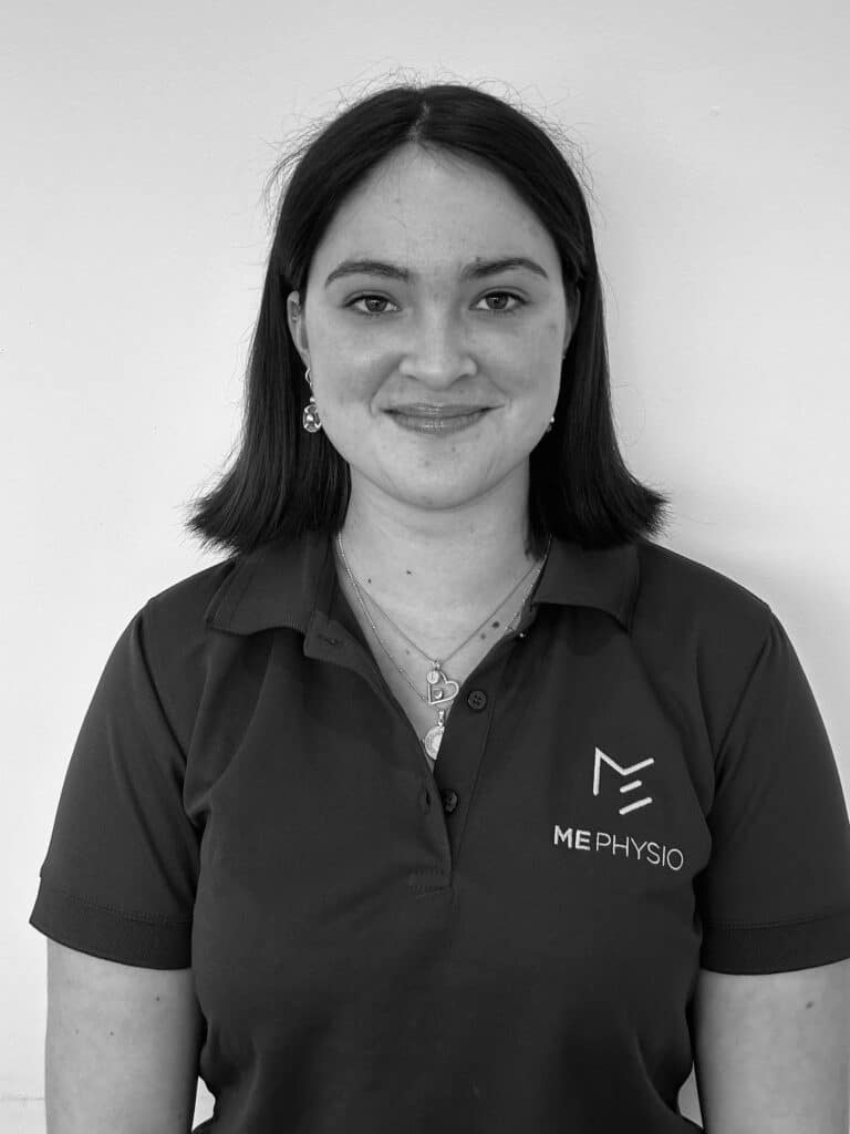 A person with shoulder-length hair is smiling at the camera while wearing a polo shirt with a "ME PHYSIO" logo on the left side. The black-and-white photo captures them standing against a plain, light-colored background, embodying the friendly professionalism of our team.