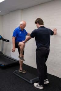 An older man in a blue shirt is balancing on a step with one knee raised, assisted by a younger man in a black shirt. They are in a gym setting with a treadmill nearby.