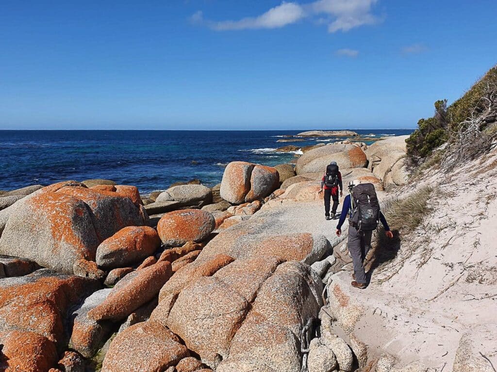 Two hikers walk along a sandy path surrounded by large, orange-tinged boulders near a sparkling blue ocean under a clear sky. Bushes and rocks line the path, creating a rugged coastal landscape.