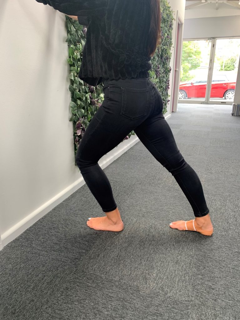 A person in black jeans and a black top does a calf stretch against a white wall, barefoot on a gray carpet&mdash;an ideal rehab move for Calf Muscle Strain. Leafy green wall decor and a window with a red car are visible in the background.