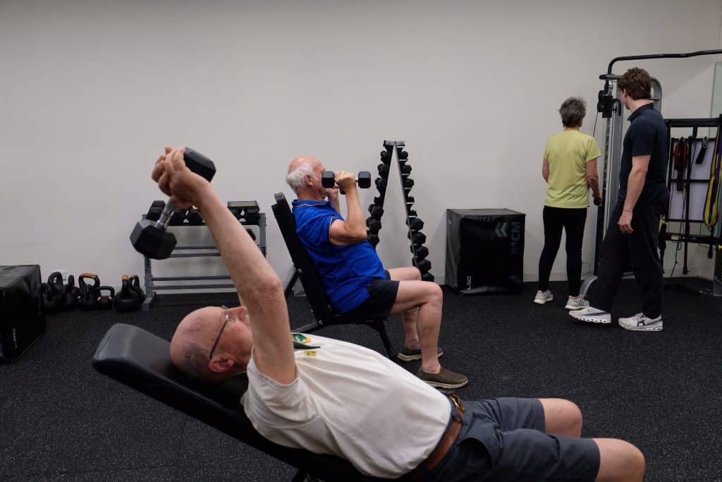 Two older men use dumbbells on benches in a gym, showcasing clinical strength; one lifts weights overhead while the other curls. In the background, a woman and younger man stand near exercise equipment beside racks of dumbbells and kettlebells.