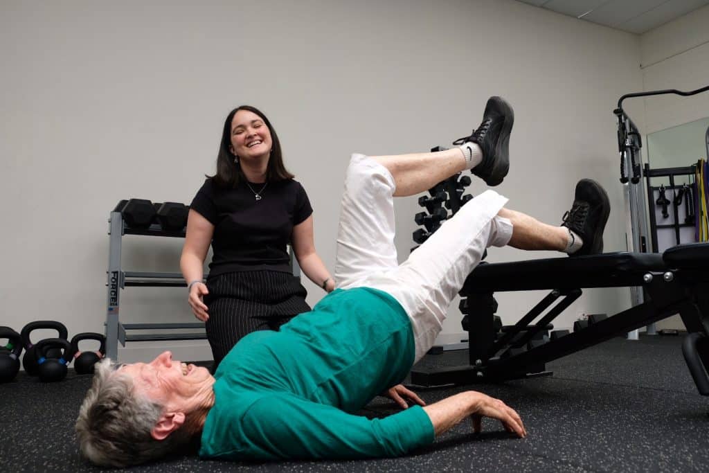 An older adult performs a leg exercise on a bench in a gym, building clinical strength while a younger woman, possibly a trainer, smiles and encourages them. Fitness equipment is visible in the background.