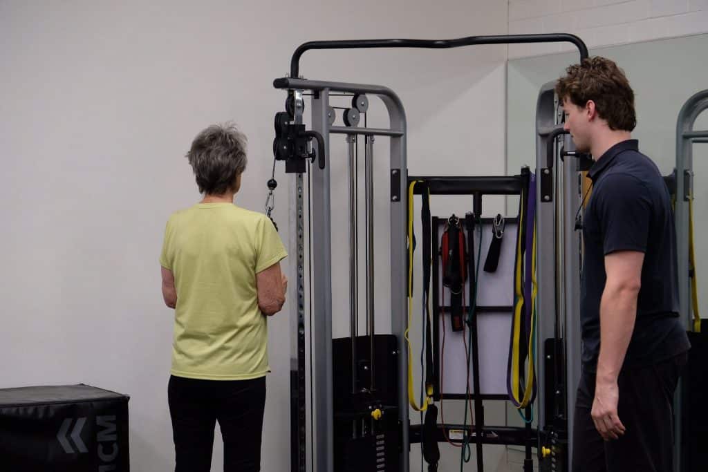 An older woman in a yellow shirt uses a cable machine at a gym, building clinical strength as a male trainer observes. Various resistance bands and gym equipment are visible in the background.