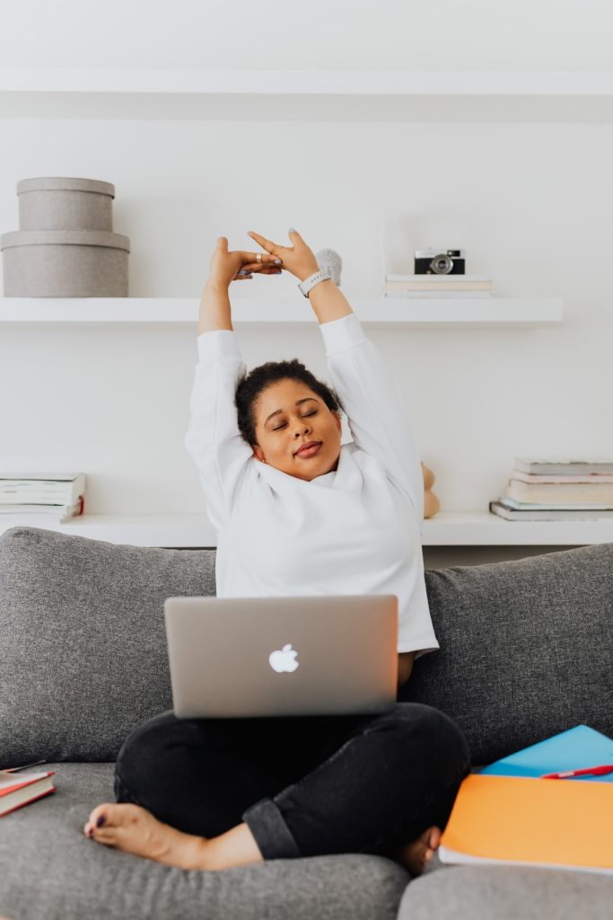 Woman sitting on a gray sofa with a laptop on her lap, stretching her arms overhead and closing her eyes&mdash;perhaps relieving back muscle strain&mdash;with shelves, books, and boxes in the background.