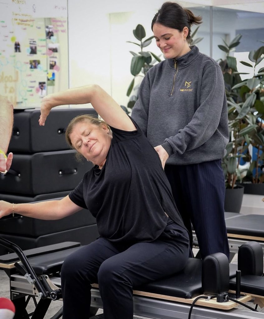 A woman in black exercises on a Pilates reformer, stretching her arm overhead&mdash;an ideal move for addressing intercostal muscle strain&mdash;while an instructor in a gray sweatshirt supports her back. They are in a bright studio with plants and equipment.