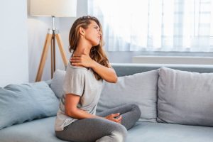 A woman sits on a couch at home in casual clothes, touching her neck and shoulder with a pained expression, suggesting she is experiencing muscle strain on neck or shoulder.