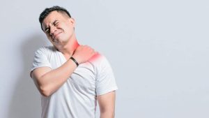 A man in a white t-shirt holds his left shoulder in pain, his face showing discomfort. A red area highlights muscle strain on neck and shoulder, indicating possible injury.