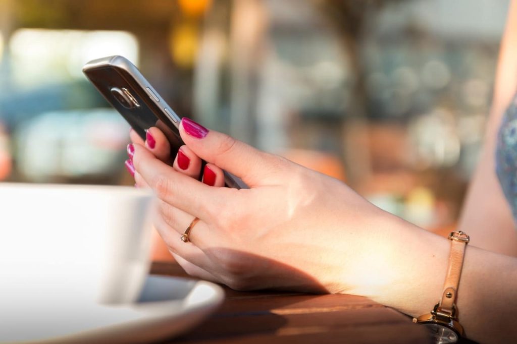 A person with painted pink nails and a gold watch, possibly adjusting their grip due to wrist pain, is holding a smartphone while sitting at an outdoor table. A white cup sits in the foreground, with the background softly blurred.