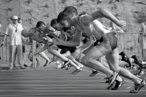 A group of male sprinters in athletic gear powerfully launch from the starting blocks at the beginning of a race on an outdoor track, captured in black and white&mdash;each pushing past limits despite risks like shin splints. Spectators and officials watch in the background.