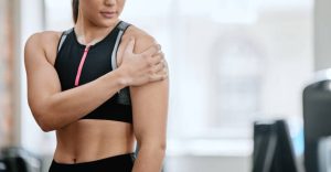 A woman in athletic wear stands indoors, holding her shoulder with one hand, suggesting rotator cuff soreness or injury. Her face is partially out of frame, with gym equipment visible in the background.