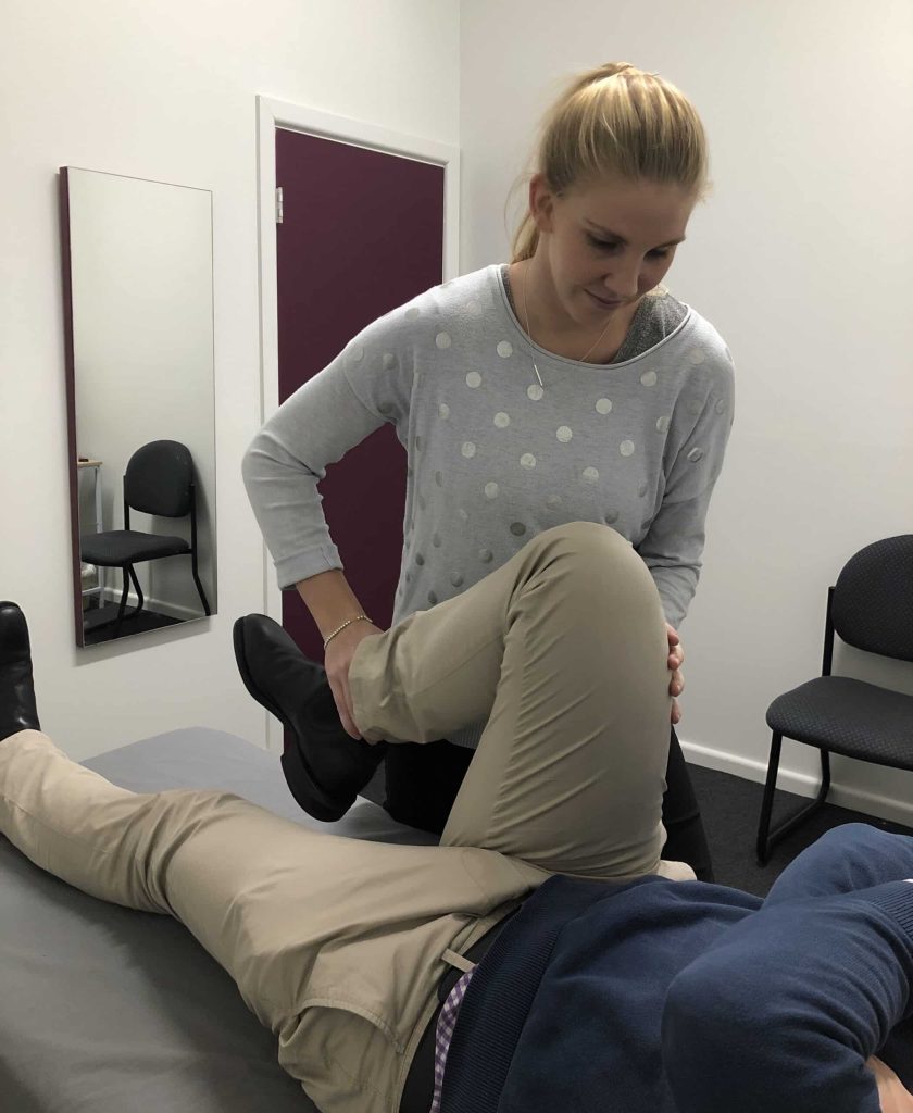 A woman performs a physical therapy exercise on a person's bent leg, helping to rehabilitate a meniscal tear while the person lies on an examination table in a clinic room.