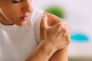 A person wearing a white sleeveless shirt touches their upper arm, appearing to check or soothe it, possibly due to shoulder pain. The background is blurred.