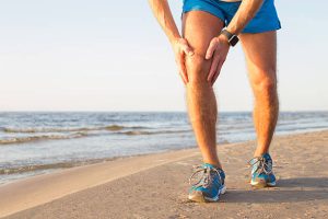 A person in blue shorts and running shoes stands on a sandy beach, holding their knee in apparent pain or discomfort&mdash;possibly due to an ACL injury&mdash;with the ocean and sky in the background.