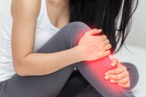 A woman in a white tank top and gray pants sits holding her lower leg, a red glow around her shin and knee indicating shin splints or injury. Her face is not visible.