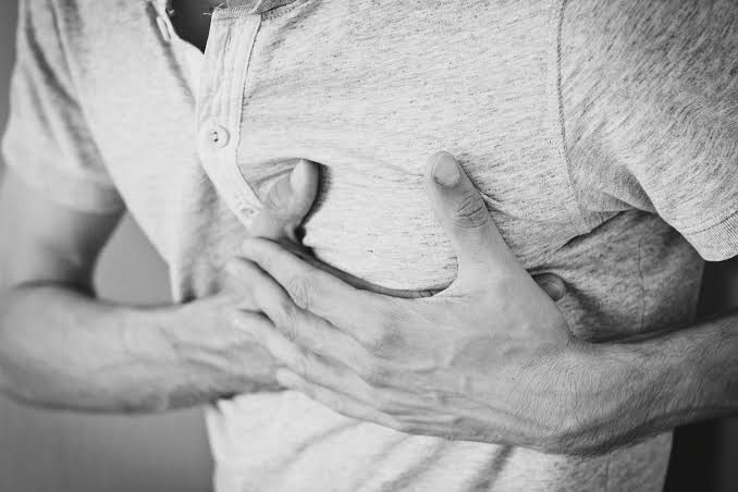 A person wearing a grey shirt clutches their chest with both hands, possibly indicating symptoms of chest pain or discomfort, which may be related to conditions such as costochondritis and could require appropriate treatment.