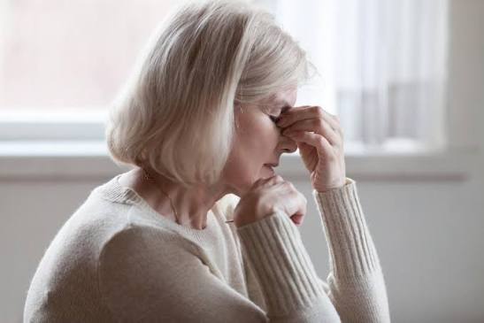An older woman sits indoors with her eyes closed, pinching the bridge of her nose and resting her elbow on a table, appearing stressed or fatigued—perhaps coping with perimenopause symptoms. Light filters through a window in the background.